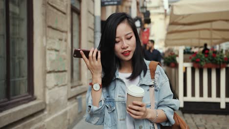 Happy-And-Atractive-Stylish-Young-Woman-Walking-The-Street-With-A-Coffee-To-Go-And-Listening-To-The-Audio-Voice-Message-On-The-Smartphone