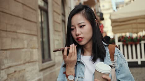 Young-Stylish-Attractive-Woman-Going-At-The-Street-Of-Touristic-City-With-A-Coffee-To-Go-In-Hand-And-Recording-A-Voice-Message-On-The-Smartphone