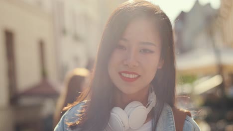 Portrait-Of-The-Beautiful-Young-Woman-With-Big-Whit-Eheadphones-On-Her-Neck-Turning-Her-Dace-And-Smiling-Cheerfully-To-The-Camera-In-Sunlight-While-Walking-As-A-Tourist