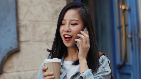 Pretty-Brunette-Young-Woman-Talking-Cheerfully-On-The-Mobile-Phone-And-Holding-A-Coffee-To-Go-In-Hand