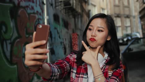 Schöne-Und-Süße-Junge-Frau-Mit-Kopfhörern-Und-Kunterbuntem-Rotem-Hemd,-Die-Fröhlich-Lächelt-Und-Vor-Der-Smartphone-Kamera-Posiert,-Während-Sie-Selfie-Fotos-An-Der-Graffity-Wand-Im-Freien-Macht