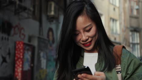 Attractive-Young-Woman-In-Stylish-Outfit-Chatting-And-Typing-On-The-Smartphone-With-Interest-And-Smile-At-The-Street-With-Graffity