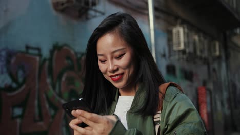 Close-Up-Of-The-Young-Joyful-Good-Looking-Woman-Walking-The-Slums-Street-With-Graffity-And-Chatting-While-Typing-Mesages-On-The-Smartphone