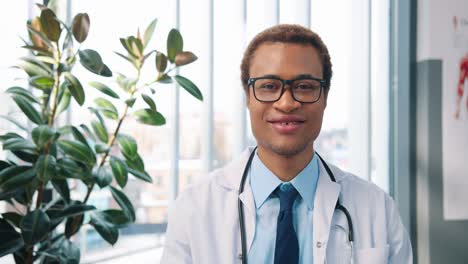 Close-Up-Portrait-Of-Happy-Handsome-Young-Male-Doctor-In-White-Coat-And-Eyeglasses-Looking-At-Camera-In-Cabinet-In-Hospital-At-Workplace,-Man-Infectionist-In-Clinic,-Health-Concept