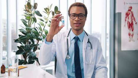 Portrait-Of-Handsome-Joyful-Young-Male-Infectionist-In-Glasses-Sitting-In-Cabinet-In-Clinic-At-Work-Speaking-On-Video-Call-On-Webcam,-Showing-Patient-New-Vaccine,-Vaccination-Concept