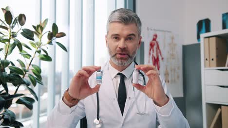 Close-Up-Portrait-Of-Happy-Handsome-Middle-Aged-Experienced-Male-Doctor-Immunologist-Looking-At-Camera-And-Speaking-On-Video-Call-With-Patient-In-Cabinet-In-Hospital,-Showing-Covid-Vaccines