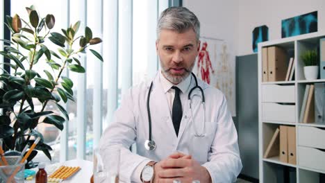 Close-Up-Portrait-Of-Handsome-Senior-Joyful-Male-Doctor-Infectionist-In-White-Coat-Sitting-In-Cabinet-In-Clinic-At-Work-Talking-On-Video-Call-Online-Through-Webcam-With-Patient-Medic-Concept