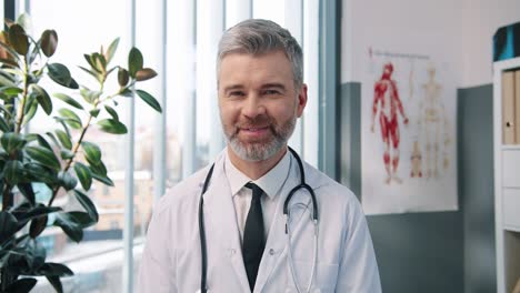 Close-Up-Portrait-Of-Handsome-Cheerful-Positive-Senior-Male-Doctor-In-White-Medical-Coat-Looking-At-Camera-And-Smiling-In-Cabinet-In-Clinic,-Man-Healthcare-Specialist,-Hospital-Concept
