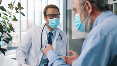 Close-Up-Of-Young-Handsome-Male-Doctor-Specialist-In-Medical-Mask-Talking-With-Old-Patient-Explaining-New-Coronavirus-Vaccine-On-Medical-Consultation,-Vaccination-Concept
