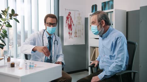 Happy-Young-Handsome-Male-Physician-Specialist-In-Medical-Mask-Talking-With-Senior-Patient-Showing-And-Explaining-Coronavirus-Vaccine-Doctor-Consultation-Vaccination-Concept