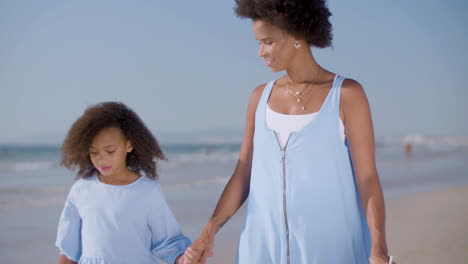 Mum-And-Daughter-Holding-Hands-And-Walking-Along-Seashore-On-A-Sunny-Day