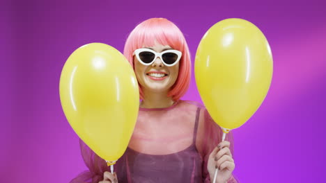 Porträtaufnahme-Einer-Schönen-Frau,-Die-Eine-Rosa-Perücke-Und-Eine-Brille-Trägt-Und-Mit-Gelben-Luftballons-Tanzt