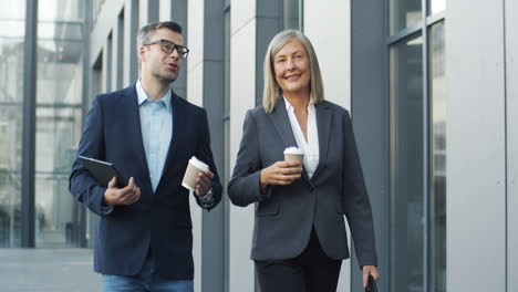 Cuc-Businessman-And-Businesswoman-Walking-The-Street-Together-With-Coffee-To-Go-In-Hands-And-Talking-Cheerfully