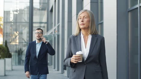 Senior-Woman-Walking-The-Street-With-Coffee-To-Go-In-Morning,-While-In-The-Background-A-Businessman-Is-Talking-On-Mobile-Phone