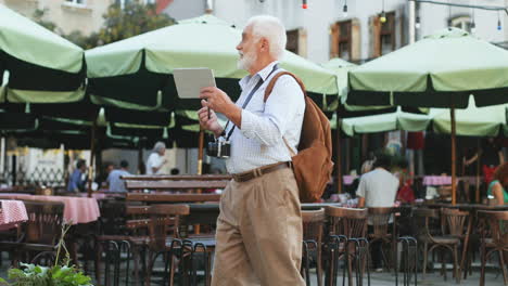 Anciano-Caminando-Como-Turista-En-Una-Terraza-De-Café-En-La-Ciudad-Y-Videochat-En-El-Dispositivo-De-Tableta