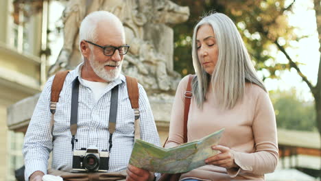 Portrait-Of-A-Couple-Of-Senior-Tourists-Sitting-On-The-Monument-In-The-Center-Of-A-City,-Talking-And-Looking-At-The-Map
