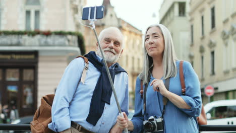 Smiling-Couple-Of-Tourists-Standing-In-The-City-Center-With-A-Smartphone-On-The-Selfie-Stick-And-Having-A-Videochat-Or-Taking-Photos