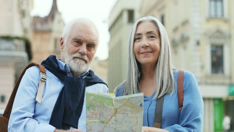 Portrait-Of-A-Senior-Couple-Of-Tourists-Standing-In-The-City-And-Looking-At-A-Map,-Then-Smiling-To-The-Camera
