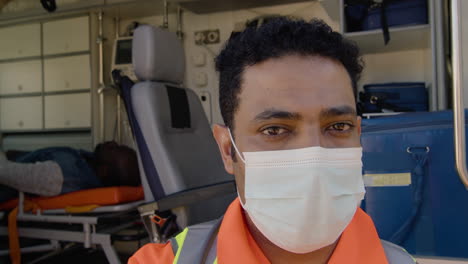 Portrait-Of-A-Latin-Male-Paramedic-With-Face-Mask-Standing-In-Front-Of-An-Ambulance-And-Looking-At-The-Camera