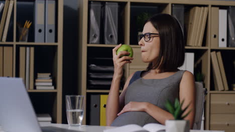 Pregnant-Woman-In-Glasses-Biting-A-Green-Apple-While-Resting-In-The-Office-At-Her-Working-Place