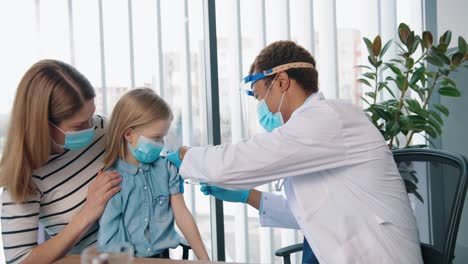 Portrait-Of-Young-Handsome-Happy-Man-Doctor-Specialist-Immunologist-In-Mask-Injecting-Cute-Small-Kid-Girl-Sitting-In-Clinic-With-Mom,-Disease-Prevention,-Vaccination-Concept
