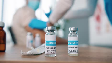 Nahaufnahme-Von-Coronavirus-impfampullen-Mit-Flüssigkeit,-Ampulle-Mit-Impfstoff-Von-Covid-19-Auf-Dem-Tisch-Im-Klinikraum,-Arzt,-Der-Im-Hintergrund-Injiziert,-Injektionsset,-Impfkonzept