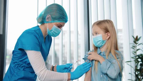 Close-Up-Portrait-Of-Cute-Little-Girl-Child-Receiving-Covid-19-Vaccine-In-Hospital,-Woman-Doctor-Or-Nurse-In-Medical-Mask-Injecting-Coronavirus-Vaccine-To-Small-Cute-Kid-In-Medical-Mask