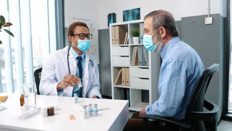 Portrait-Of-Pensioner-Male-Patient-In-Medical-Mask-Sitting-In-Hospital-Cabinet-Listening-Young-Experienced-Professional-Doctor-Showing-And-Discussing-Vaccines-On-Consultation