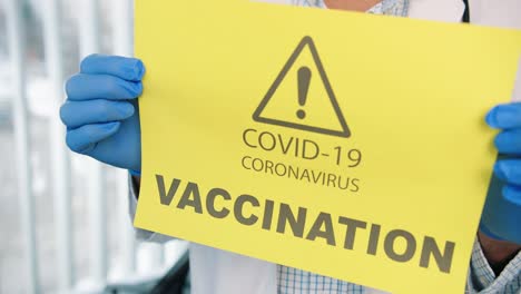 Nahaufnahme-Von-Personenhänden-In-Schutzhandschuhen,-Die-Eine-Papierkarte-Mit-Ankündigung-über-Coronavirus-Impfkampagne,-Covid-19-Impfstoff,-Aufmerksamkeit,-Gesundheitsbenachrichtigung-Halten