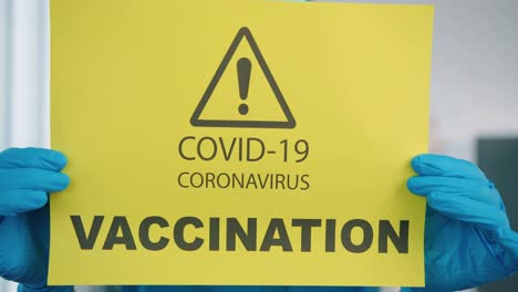 Close-Up-Of-Young-Pretty-Female-Healthcare-Worker-Medic-In-Protective-Suit-And-Medical-Mask-Holding-In-Hands-Yellow-Paper-Card-With-Notification-About-Covid-19-Coronavirus-Vaccination