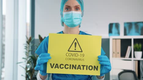 Primer-Plano-De-Una-Hermosa-Doctora-Especialista-En-Guantes-Protectores-Y-Máscara-Médica-Sosteniendo-En-Las-Manos-Una-Tarjeta-De-Papel-Amarillo-Que-Muestra-Una-Notificación-Sobre-La-Vacunación-Covid-19,-Pandemia-De-Coronavirus