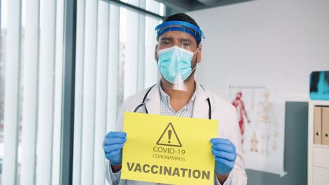 Close-Up-Portrait-Of-Young-Man-Doctor-In-Protective-Shield-And-Gloves-Holding-In-Hands-Yellow-Paper-Card-Showing-Notification-About-Covid-19-Vaccination,-Coronavirus-Pandemic