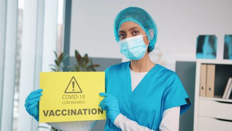Retrato-De-Una-Joven-Enfermera-Especialista-Hindú-Con-Guantes-Protectores-Y-Máscara-Médica-Sosteniendo-En-Las-Manos-Una-Tarjeta-De-Papel-Amarilla-Que-Muestra-Un-Anuncio-Sobre-La-Vacunación-Covid,-La-Vacuna-Contra-El-Coronavirus