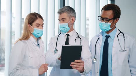 Portrait-Of-Healthcare-Colleagues-Multi-Ethnic-Female-And-Male-Doctors-In-Medical-Masks-Standing-In-Clinic-Chatting-Discussing-Patient-Analysis-Results-And-Treatment,-Typing-On-Tablet-Pandemic-Concept