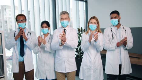 Portrait-Of-Experienced-Happy-Positive-Different-Mixed-Races-Healthcare-Workers-Doctors-And-Physicians-In-Medical-Masks-Stand-In-Clinic-Clapping-Hands-And-Applauding-Quarantine-Concept-Team-Colleagues