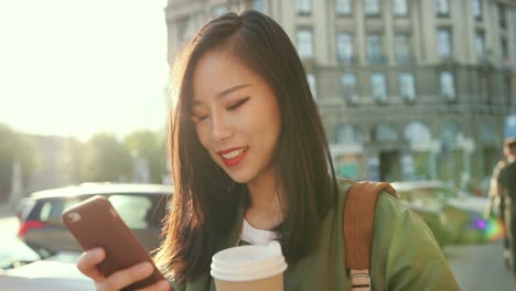 Close-Up-Of-The-Smiled-Young-Beautiful-Woman-Typing-A-Message-On-The-Smartphone-And-Laughing-At-The-Noisy-Street-Of-The-Town