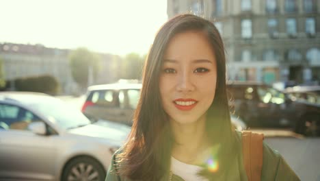 Portrait-Shot-Of-The-Charming-Youung-Stylish-Woman-Smiling-Happily-To-The-Camera-And-Fixing-Her-Hair-With-A-Hand-At-The-Noisy-Street-With-Traffic