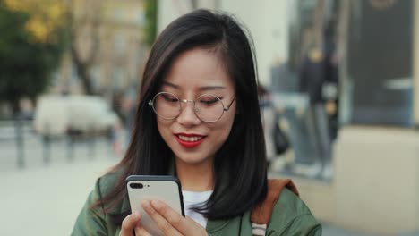 Close-Up-Of-The-Cheerful-Young-Woman-In-Glasses-Tapping-And-Texting-A-Message-On-The-Smartphone-Device-In-Hands