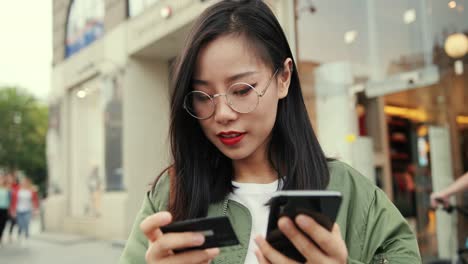 Close-Up-Of-The-Young-Beautiful-Girl-In-Glasses-Standing-At-The-Street-With-A-Credit-Card-In-Hand-And-Shopping-Online-On-The-Smartphone