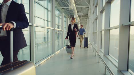 Business-People-Walking-The-Airport-Passage-With-Suitcases-On-Wheels-To-The-Plane-And-Speking-On-Phone