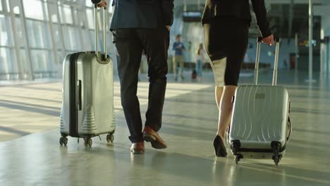 Back-View-On-The-Businessman-And-Businesswoman-Walking-The-Airport-Hall-And-Carrying-Their-Suitcases-On-The-Wheels-Before-The-Departure-To-The-Working-Trip