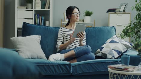 Young-Good-Looking-Woman-In-Glasses-Using-Tablet-Device-And-Thinking-While-Sitting-On-The-Blue-Sofa-At-Home