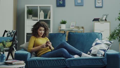 Attractive-Young-Curly-Lady-Sitting-On-The-Sofa-And-Resting-While-Tapping,-Scrolling-And-Texting-On-The-Smartphone-At-Home