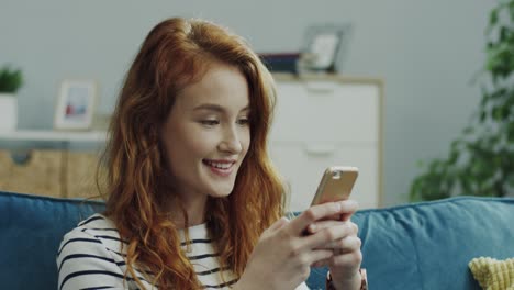 Young-Cheerful-Girl-With-Red-Hair-Tapping-And-Typing-On-The-Smartphone-While-Sitting-On-The-Couch-At-Home