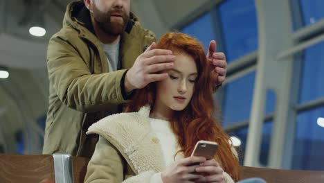 Young-Attractive-Girl-With-Red-Hair-Sitting-In-The-Airport-And-Waiting-For-Her-Departure-While-Texting-On-The-Smartphone,-Then-Her-Boyfriend-Or-Male-Friend-Coming-From-Behind-And-Closing-Her-Eyes