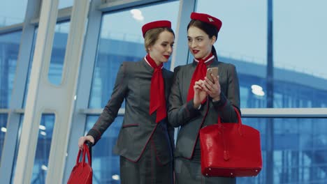 Zwei-Schöne-Junge-Stewardessen,-Die-Durch-Die-Flughafenpassage-Gehen,-Sich-Unterhalten-Und-Eine-Der-Anderen-Etwas-Auf-Dem-Smartphone-Zeigt