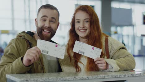 Portrait-Of-The-Young-Cheerful-Couple-Standing-At-The-Checking-Desk-At-The-Airport-With-Tickets-In-Hands-And-Smiling-To-The-Camera