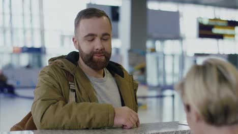 Close-Up-Of-The-Young-Smiled-Guy-With-Beard-Coming-To-The-Checking-Desk-At-The-Airport,-Giving-His-Passport-And-Ticket
