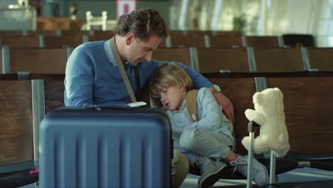 Vater-Mit-Seinem-Kleinen-Süßen,-Verschlafenen-Sohn,-Der-Mit-Koffern-Am-Flughafen-Sitzt-Und-Auf-Ihren-Flug-Wartet