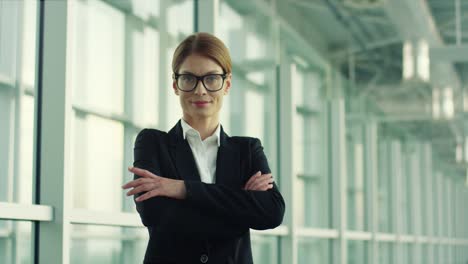 Portrait-Of-The-Beautiful-Businesswoman-In-Glasses-Standing-In-The-Big-Hall-Like-Of-Airport,-Turning-To-The-Camera,-Smiling-And-Crossing-Her-Hands-In-Front-Of-Her